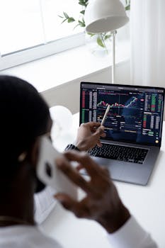 A businessman using a smartphone while analyzing stock market graphs on a laptop indoors.