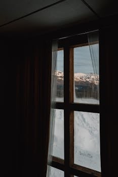 Snowy mountain view through a rustic cabin window evokes tranquility and solitude.