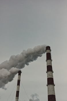 Two tall industrial chimneys releasing white smoke into a cloudy gray sky, illustrating air pollution.